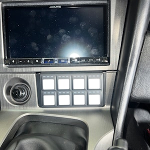 May include: A close-up of a car's dashboard with a black Alpine car stereo and a panel of 12 white toggle switches.