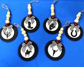 May include: Six rustic, western-themed ornaments. Each ornament features a black circular base with a white center displaying a cowboy boot, cactus, cowboy hat, horseshoe, bull skull, and a cowboy on a horse. They have a gingham bow and wooden bead accents.