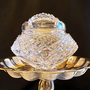 Puede incluir: Una fuente de servir plateada vintage y ornamentada con un tarro y tapa de cristal transparente tallado en diamante. La fuente tiene un borde festoneado y un tallo retorcido, que descansa sobre una base circular. El tarro está diseñado para guardar objetos pequeños.