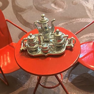 May include: A miniature red metal table and chairs set with a silver tea set on the table. The tea set includes a teapot, creamer, sugar bowl, and two cups.