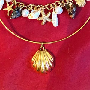 May include: Gold-toned necklace featuring seashell charms, pearls, and a large shell pendant. The necklace has a circular design with a chain and clasp closure. Two seashells are placed above the necklace on a red background.