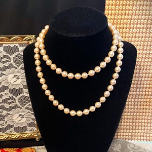 May include: A double-strand necklace featuring uniform, cream-colored round pearls. The necklace is displayed on a black velvet bust, showcasing the smooth, lustrous surface of each individual pearl arranged in two distinct, layered circular loops.