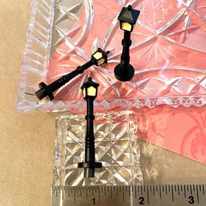 May include: Three miniature black street lamps with yellow light bulbs. The lamps are on a clear glass surface with a pink background.