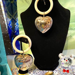 May include: A silver heart-shaped pendant with a white ring and a safety pin, displayed on a black mannequin. A second pendant is visible. A small crystal bear with a red bow tie and a gold spoon are also present.