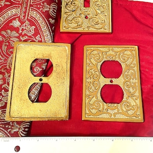 May include: Three ornate, gold-colored electrical switch plates. One is for a single switch, and the other two are for outlets. The plates have a textured, decorative design. They are displayed on a red fabric background.