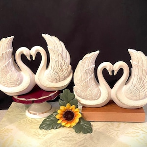 Puede incluir: Dos figuras de cisnes blancos con cuellos en forma de corazón y alas extendidas. Un par descansa sobre un pedestal rojo y blanco, el otro sobre una base de madera. Un girasol amarillo con hojas verdes está en primer plano.