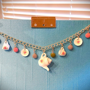 May include: A gold chain charm bracelet with various teacup and saucer charms, including a teapot charm. The charms are white with floral designs and pink and green accents.