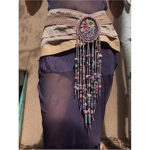 Puede incluir: Un cinturón artesanal con base de cuero beige y un adorno ovalado central. El óvalo está adornado con bordados coloridos y flecos de cuentas en tonos rosa, azul y verde. El cinturón se lleva sobre un vestido morado transparente.