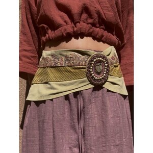May include: A wide, beige belt with a decorative oval buckle. The buckle features a heart-shaped stone surrounded by small beads. The belt has a textured, layered design with olive green and patterned fabric.