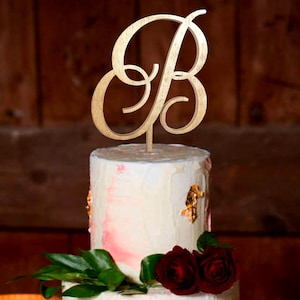 Könnte beinhalten: Ein goldfarbener Kuchenaufsatz aus Holz in Form des Buchstabens "B" sitzt auf einem weißen Kuchen mit roten Akzenten und goldenen Blattdetails. Der Kuchen ist mit roten Rosen und grünen Blättern verziert.