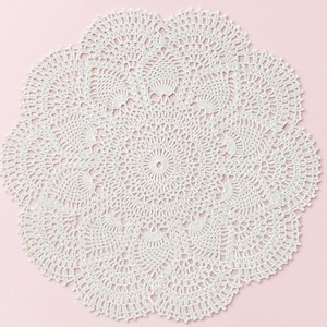 May include: A white, circular doily with a detailed, openwork crochet design. The doily features a central circular pattern surrounded by repeating floral-like motifs. The edges are scalloped, adding a decorative touch. The background is a soft pink.