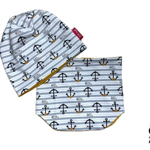 Könnte beinhalten: Ein passendes Mütze- und Halstuch-Set. Das Set hat ein maritimes Design mit grauen und weißen Streifen, schwarzen Ankern und den Wörtern "MOIN" und "AHOL". Die Innenseite der Mütze und des Halstuchs ist einfarbig gelb.