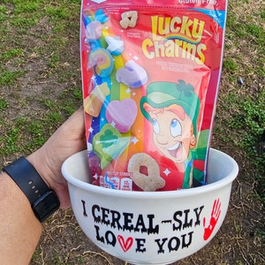 May include: A white ceramic bowl with the text "I Cereal-sly Love You" in black and red. The bowl contains a box of Lucky Charms cereal, a popular breakfast cereal with frosted oat cereal and marshmallow shapes. The box is red and features a cartoon leprechaun.