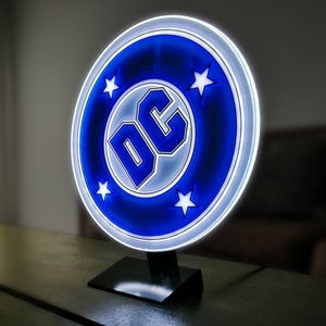 Könnte beinhalten: Eine blaue und weiße DC Comics Logo-Lampe mit fünf weißen Sternen. Die Lampe steht auf einem schwarzen Sockel.