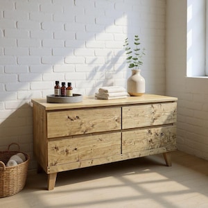 May include: A light-coloured wooden chest of drawers with four drawers, showing a natural wood grain finish. The chest is topped with a grey tray holding bottles, folded towels, and a vase with greenery. A wicker basket sits on the floor.