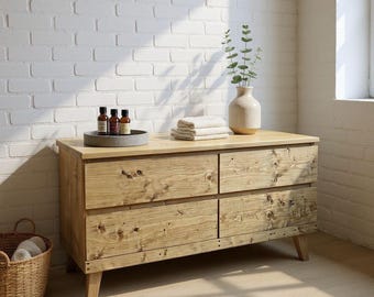 May include: A light-colored wooden dresser with four drawers, featuring a natural wood grain finish. The dresser is topped with a gray tray holding bottles, folded towels, and a vase with greenery. A wicker basket sits on the floor.