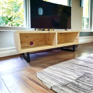 May include: A wooden TV stand with two open shelves and black metal legs. The stand is made of natural wood with a light brown finish. The stand is sitting on a light brown wood floor.