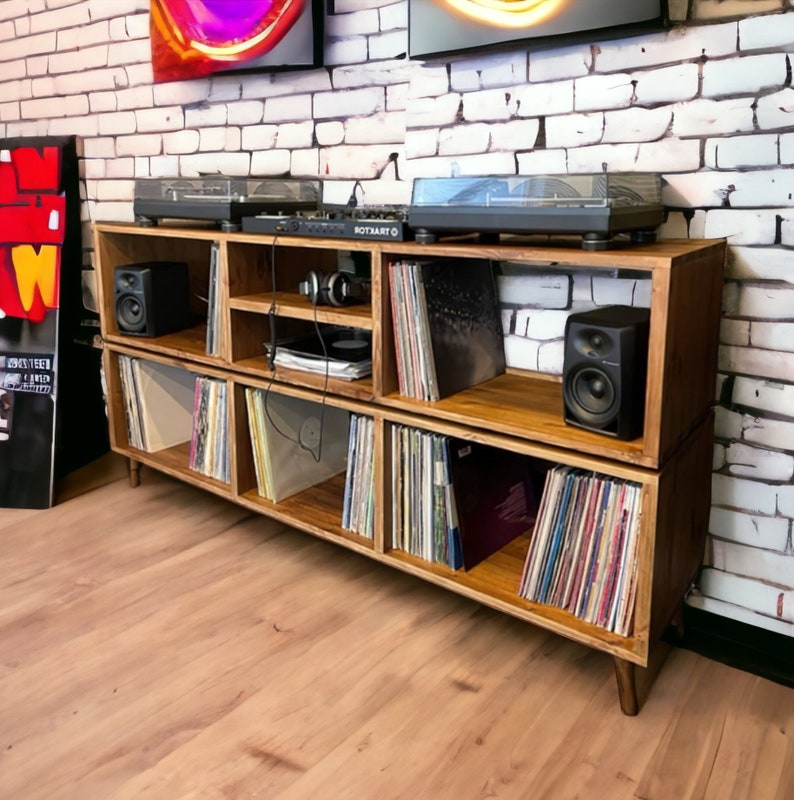 May include: A wooden record player stand with two shelves, each with multiple compartments for storing vinyl records. The stand has a dark wood finish and is decorated with a record player and speakers.
