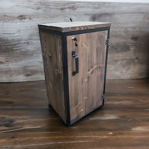 May include: A small wooden cabinet with a black metal frame and door. The cabinet has a keyhole and a black metal handle. The wood is a dark brown color.