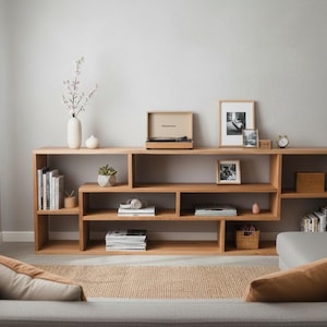 May include: A light-coloured wooden bookshelf with multiple compartments, holding books, a record player, and decorative items. A plant and a vase with flowers are also visible. The room has a neutral colour palette.