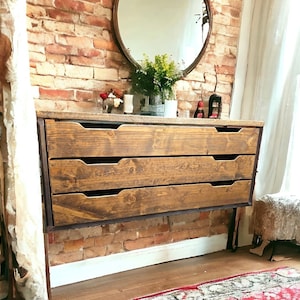 May include: A rustic wooden dresser with three drawers, featuring a dark brown stain and a black metal frame. The dresser is mounted to the wall and has a minimalist design.