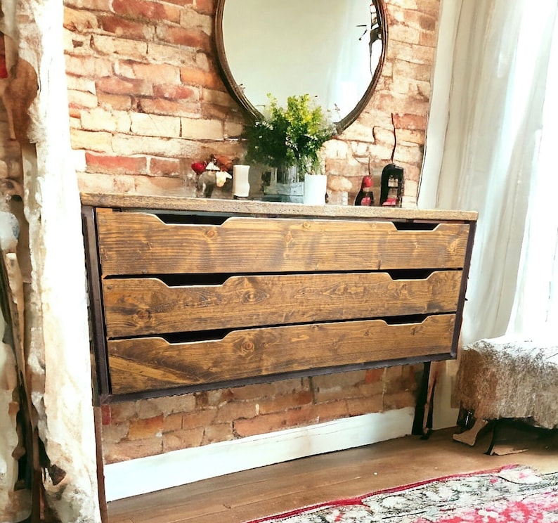 May include: A rustic wooden dresser with three drawers, featuring a dark brown stain and a black metal frame. The dresser is mounted to the wall and has a minimalist design.