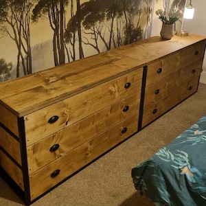 May include: Two wooden chest of drawers with black metal accents. The drawers have black knobs. The chest of drawers is in front of a wall with a forest wallpaper.