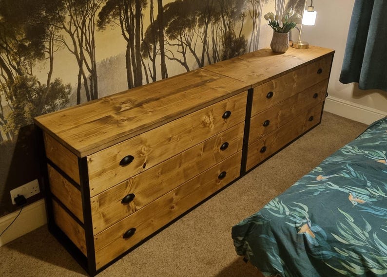 May include: Two wooden chest of drawers with black metal accents. The drawers have black knobs. The chest of drawers is in front of a wall with a forest wallpaper.