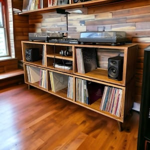 May include: A wooden record player stand with multiple shelves, featuring a turntable and speakers. The stand is made of light brown wood and has a rustic look. The shelves are filled with vinyl records.