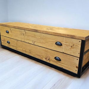 May include: A long wooden chest with two drawers and black metal accents. The chest has a natural wood finish and black metal drawer pulls.