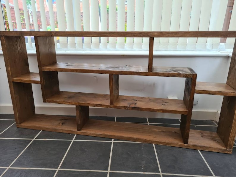 May include: A rustic wooden shelf unit with a unique asymmetrical design. The shelf has multiple compartments of varying sizes, perfect for displaying books, plants, or decorative items.
