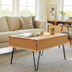 Op de afbeelding: Een licht houten salontafel met een rechthoekig blad en zwarte haarspeldpoten. De tafel heeft een lade, een boek, een mok en een kleine plant in een pot staan erop. De tafel staat in een woonkamer.