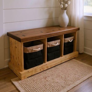 Peut inclure: Banc de rangement en bois avec trois compartiments, chacun contenant un panier en tissu noir avec un dessus beige et des poignées en corde. Le banc a un dessus marron foncé et repose sur une base marron clair. Un tapis beige est devant le banc.
