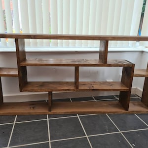 May include: A rustic wooden shelf unit with a unique asymmetrical design. The shelf has multiple compartments of varying sizes, perfect for displaying books, plants, or decorative items.