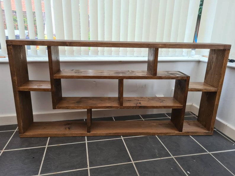 May include: A rustic wooden shelf unit with a unique asymmetrical design. The shelf has multiple compartments of varying sizes, perfect for displaying books, plants, or decorative items.