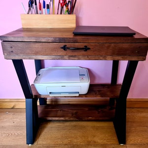 Puede incluir: Un escritorio de madera marrón oscuro con un cajón y un estante inferior. El escritorio tiene patas de metal negro y una impresora blanca en el estante inferior.