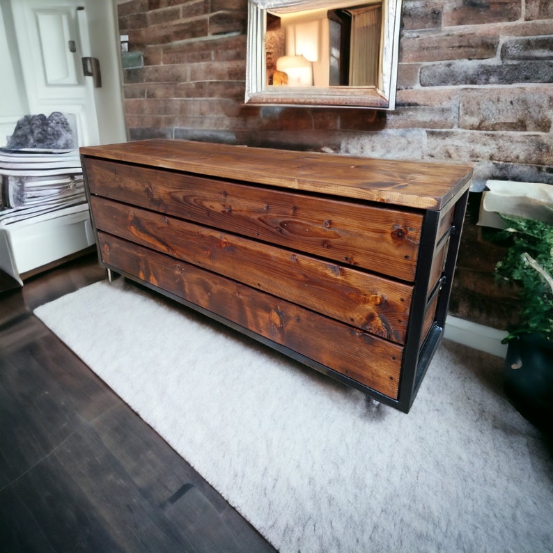 May include: A rustic wooden dresser with a black metal frame and three drawers. The dresser is sitting on a white rug in front of a brick wall.