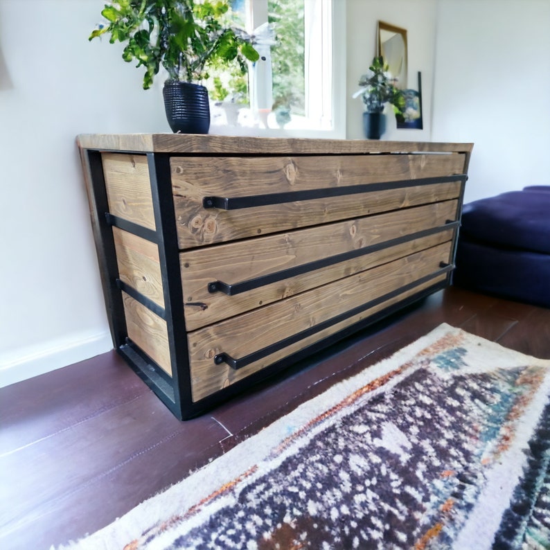 May include: A three-drawer dresser with a rustic wood finish and black metal frame. The drawers have black metal handles and are set on a dark brown floor.