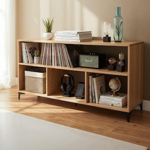 Peut inclure: Une étagère en bois de chêne clair avec des pieds en métal noir. Les étagères présentent des disques vinyles, des livres, un haut-parleur, un casque, un petit globe et des objets décoratifs. Un panier tressé et une plante en pot sont également visibles.