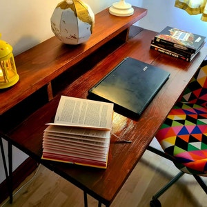 Könnte beinhalten: Ein Holzschreibtisch mit einem Laptop, einem Buch, einem Globus, einer gelben Laterne und einem Stuhl mit farbenfrohem geometrischem Muster. Der Schreibtisch hat ein Regal und schwarze Haarnadelbeine.