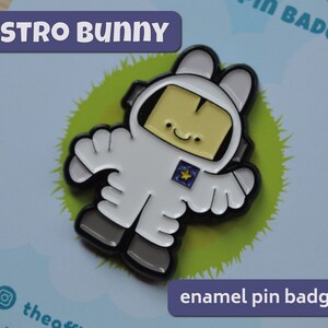 May include: A white enamel pin badge featuring a cartoon bunny dressed as an astronaut. The bunny is wearing a white spacesuit with a black outline and a yellow star on the chest. The bunny has a black outline and a yellow face with a small smile. The text "ASTRO BUNNY" is written in purple letters above the bunny. The text "enamel pin badge" is written in purple letters below the bunny.