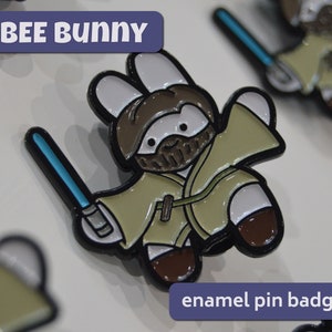 Könnte beinhalten: Ein braunes und weißes Emaille-Anstecknadel mit einem Hasen, der als Jedi-Ritter verkleidet ist und ein blaues Lichtschwert hält. Die Anstecknadel trägt die Aufschrift "Obee Bunny" und "Emaille-Anstecknadel".