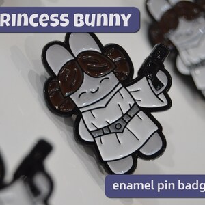 May include: Princess Bunny enamel pin badge featuring a white bunny dressed as Princess Leia with a black blaster in her hand. The bunny has brown hair and a white dress with a black belt.