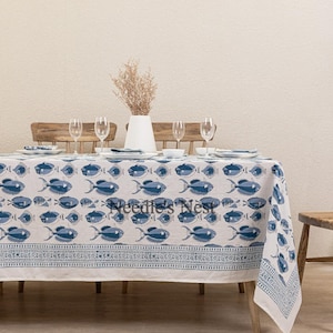 Puede incluir: Mantel blanco con un patrón de peces azules. El mantel está puesto sobre una mesa de madera con seis vasos y un jarrón con flores secas.