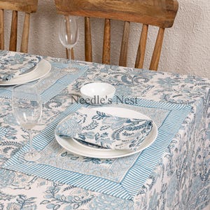 May include: A white tablecloth with a blue floral pattern and matching napkins and plates. The table is set with wine glasses and wooden chairs. The tablecloth has a striped border and the text "Needle's Nest".