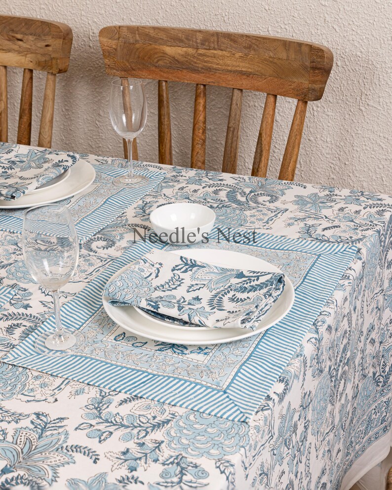 May include: A white tablecloth with a blue floral pattern and matching napkins and plates. The table is set with wine glasses and wooden chairs. The tablecloth has a striped border and the text "Needle's Nest".