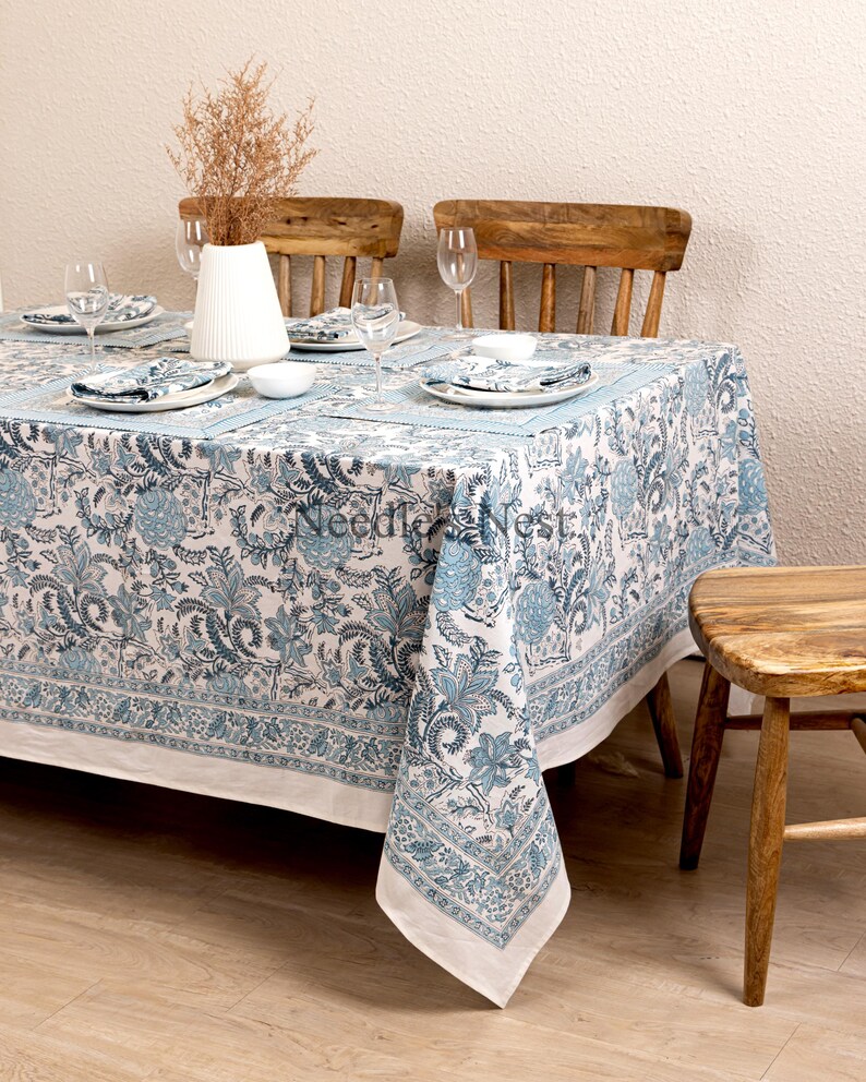 May include: A rectangular tablecloth with a blue floral pattern, set on a wooden table. The tablecloth has a white border and is paired with matching napkins and placemats. The table is set with plates, bowls, and wine glasses.