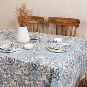 May include: A dining table set with a white and blue floral patterned tablecloth, matching napkins, and white plates. The table is set for a meal, with a white vase holding dried flowers and wooden chairs in the background.