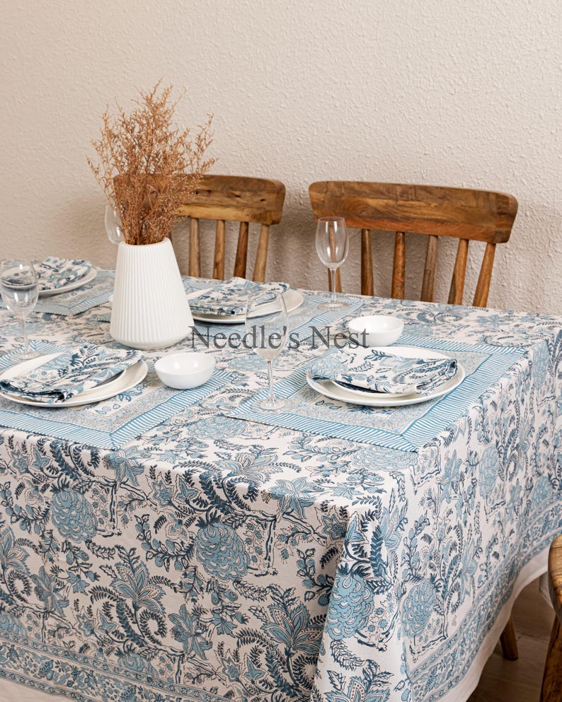 May include: A dining table set with a white and blue floral patterned tablecloth, matching napkins, and white plates. The table is set for a meal, with a white vase holding dried flowers and wooden chairs in the background.