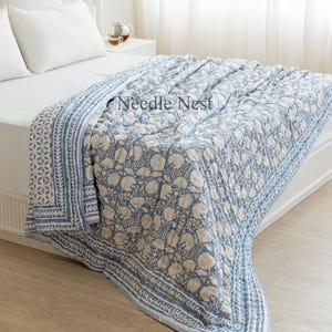 Peut inclure: Une couette à motif floral bleu et blanc avec une bordure blanche et un fond blanc. La couette est pliée sur un lit avec des oreillers blancs et une tête de lit blanche. Le texte "Needle Nest" est visible sur la couette.